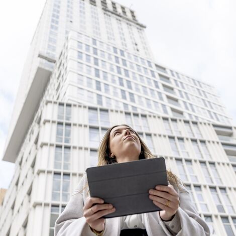 Blonde vrouw met iPad kijkt omhoog naar wit flatgebouw.
