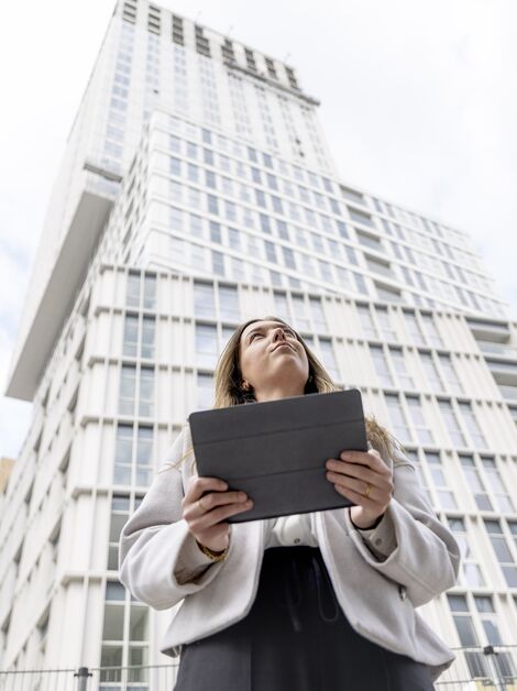 Blonde vrouw met iPad kijkt omhoog naar wit flatgebouw.