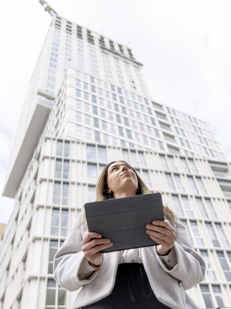 Blonde vrouw met iPad kijkt omhoog naar wit flatgebouw.